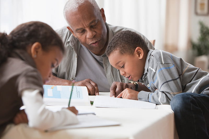Grandfather sitting at table helping grandchildren learn and write in notebooks