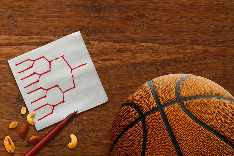stock image of a basketball and an elimination bracket drawn on a napkin on top of a table with a pen and some nuts