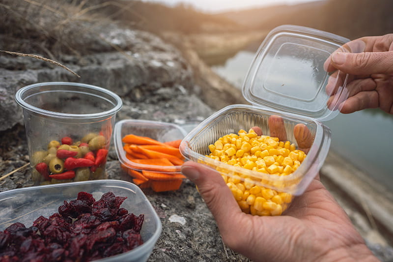 close up of hands holding a reusable container of snacks near other containers of snacks outdoors near a pond