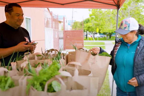 A woman is picking up a paper grocery bag of fresh produce at a Growing Together stand.