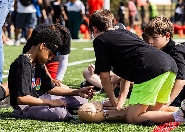 Young boys gathered on a sports field practicing CPR techniques on a manikin