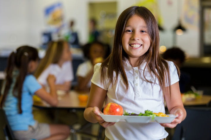 Girl in an elementary school cafeteria holding a lunch tray. 