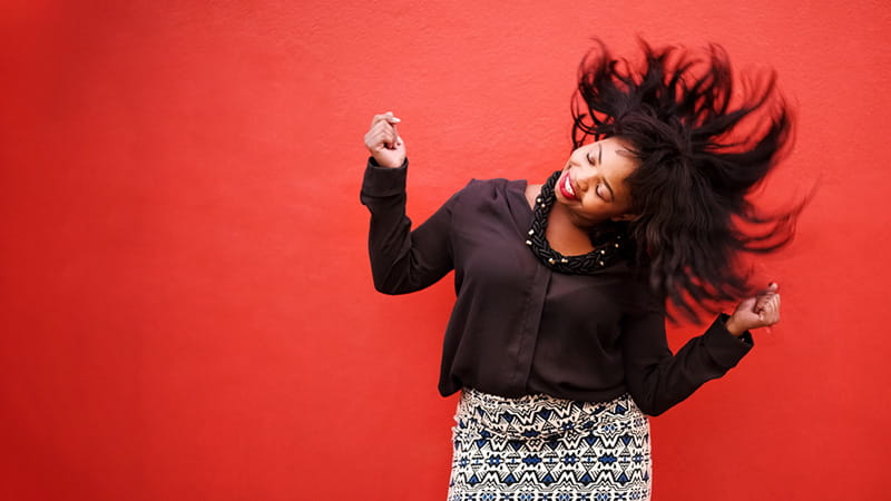 A woman is dancing and shaking her head and hair in front of a red background.