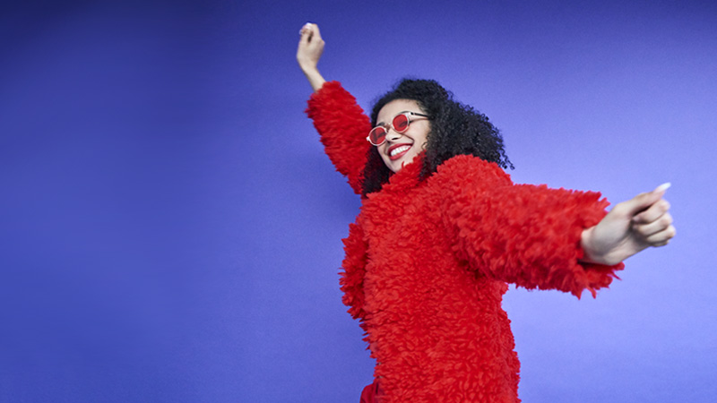 Happy young woman in a furry red coat and trendy glasses dancing on a purple background