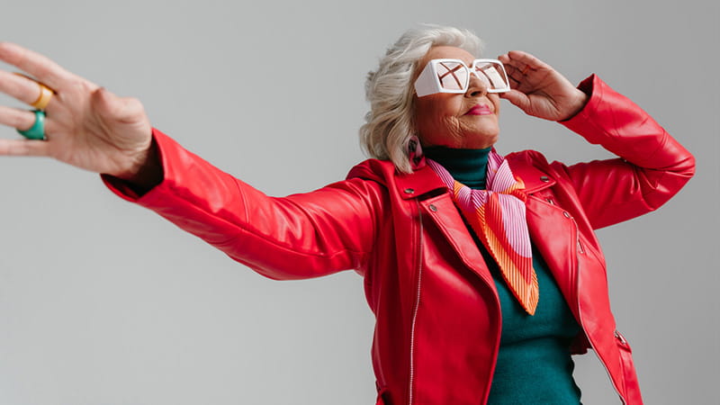 Cool older woman in a red leather jacket and trendy eyeglasses dancing on a grey background