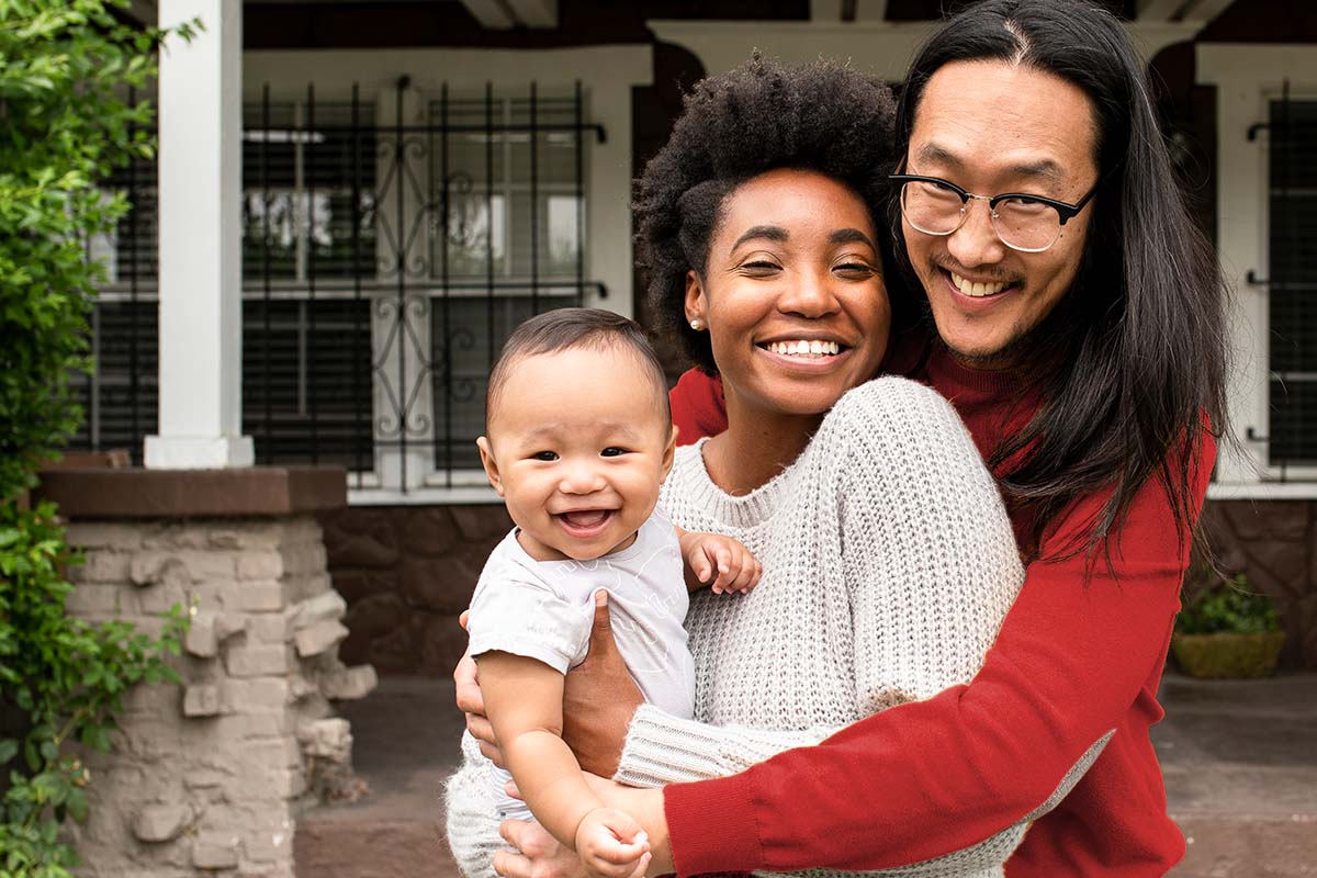 a couple smiling and hugging, holding a baby in front of a home
