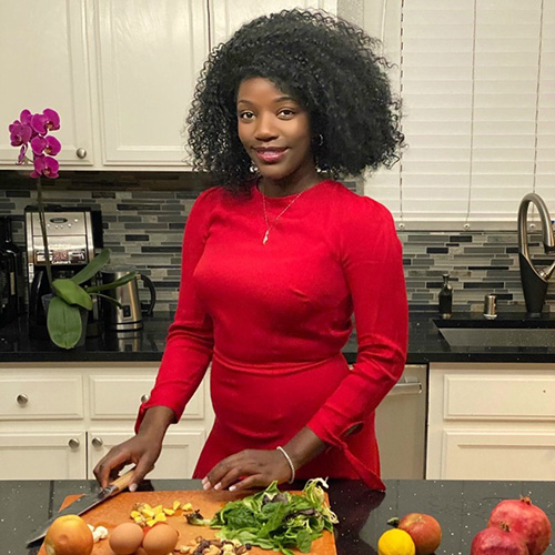 Woman in her kitchen, smiling in a red dress, chopping healthy fruits and vegetables