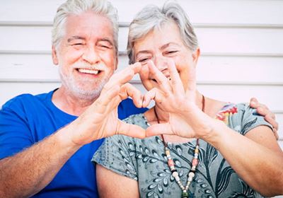 An older couple sitting together and smiling while holding their hands up to make a heart shape