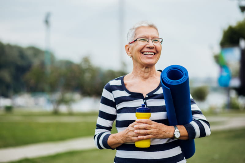 smiling older woman walking outside, carrying water bottle and yoga mat