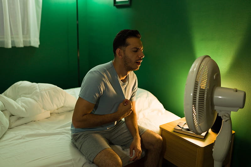 overheated man sitting on his bed facing into a floor fan, trying to cool down