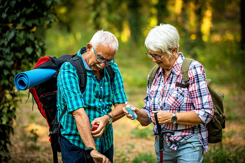 an older couple wearing backpacks stop hiking in the woods to scratch at mosquito bites and apply bug spray