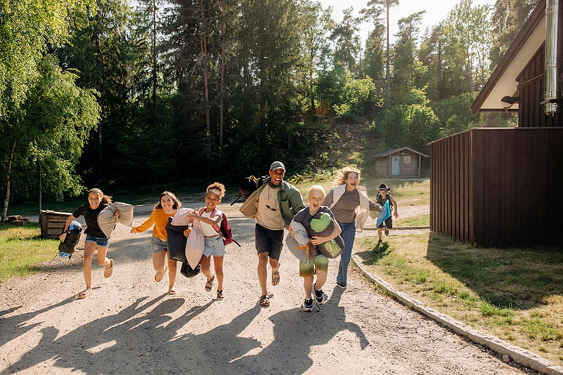 group of smiling kids carrying sleeping bags, pillows, and backpacks running with parents into a sunny campground