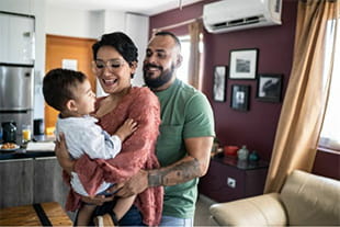 mom, dad, and toddler son smiling in living room. the mom is holding the son and dad is hugging both of them.