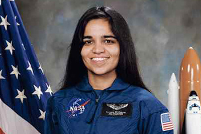 A professional portrait of Kalpana Chawla in her blue uniform with an American flag and shuttle model in the background