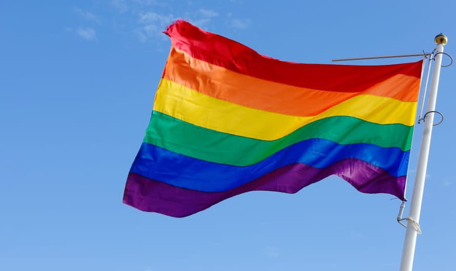 A rainbow flag flying on a flagpole against a blue sky.