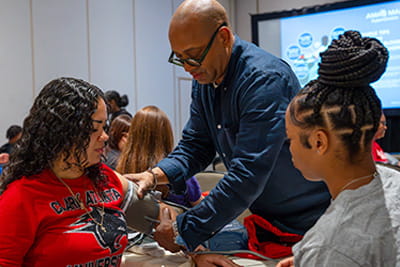 A male instructor is teaching the appropriate way to apply a blood pressure cuff to two female American Heart Association Undergraduate Scholars.