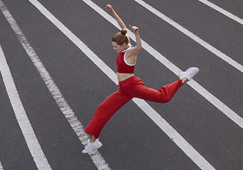 A woman in red athletic apparel is leaping over white painted lines on an asphalt surface.