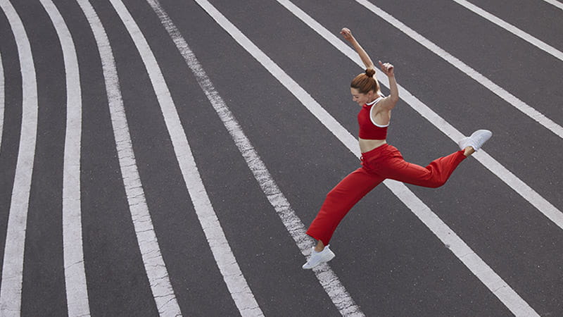 A woman in red athletic apparel is leaping over white painted lines on an asphalt surface.