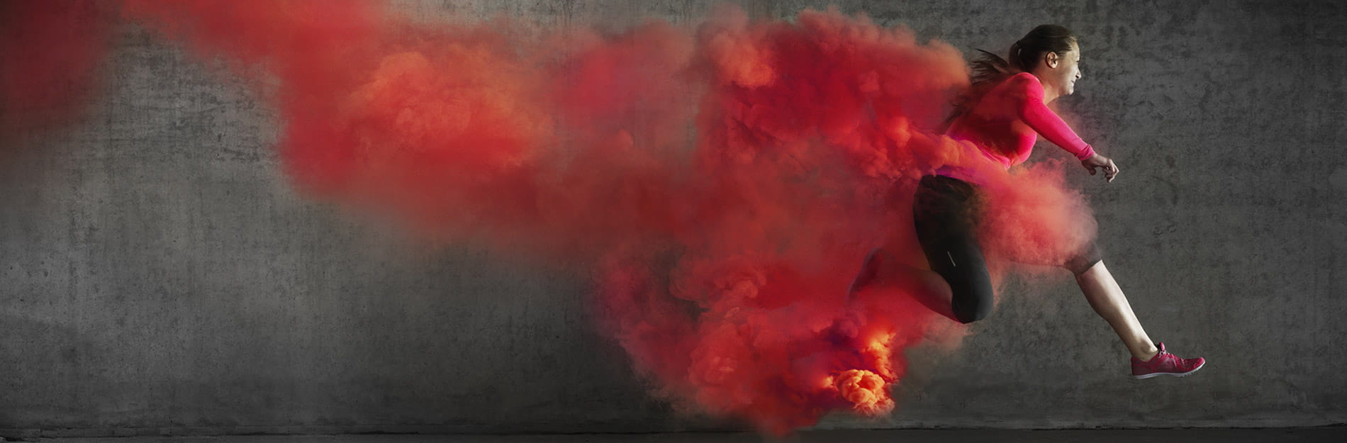 A woman in athletic apparel is running through a trail of red smoke on a grey background.