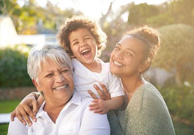 A mother, a grandmother, and a young child smiling and posing together outside in the sunshine