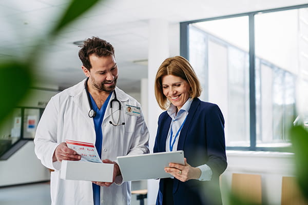 A doctor in a white coat with a person in business attire smiling and looking at a tablet together